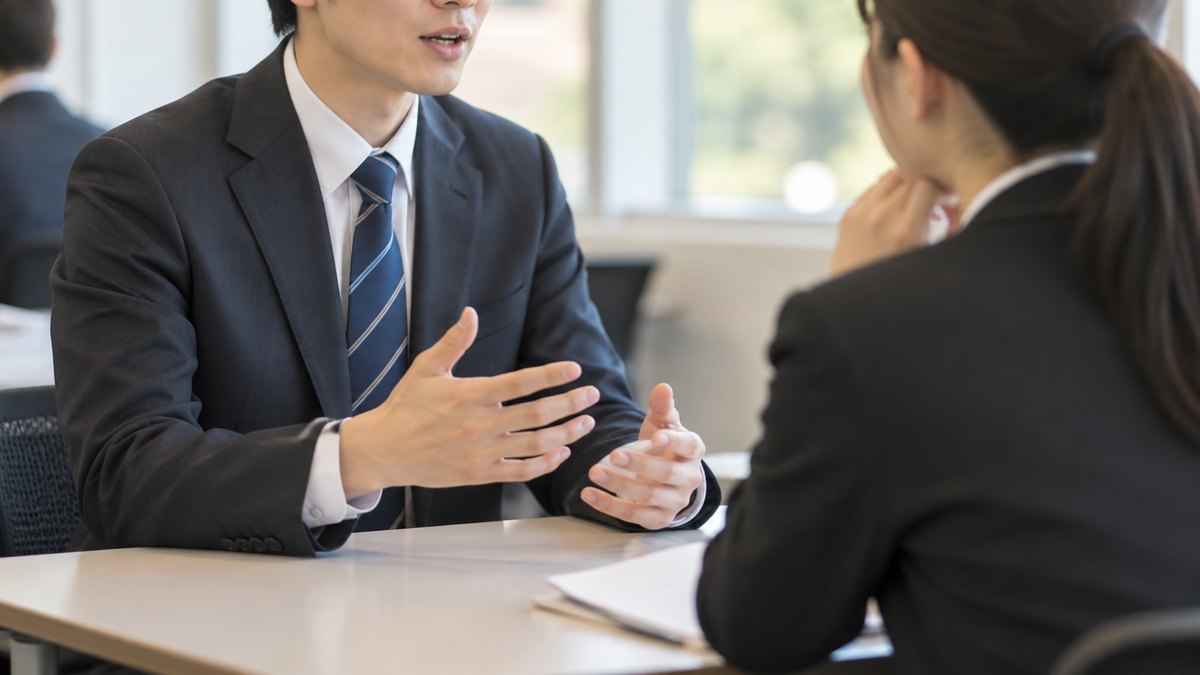 午後、研修室の一角で、スーツ姿の20代の新入社員2人が向かい合って座り、1人が話し、もう1人がうなずきながら聞くロールプレイをしている近景。顔ははっきり写らない。文字は無い