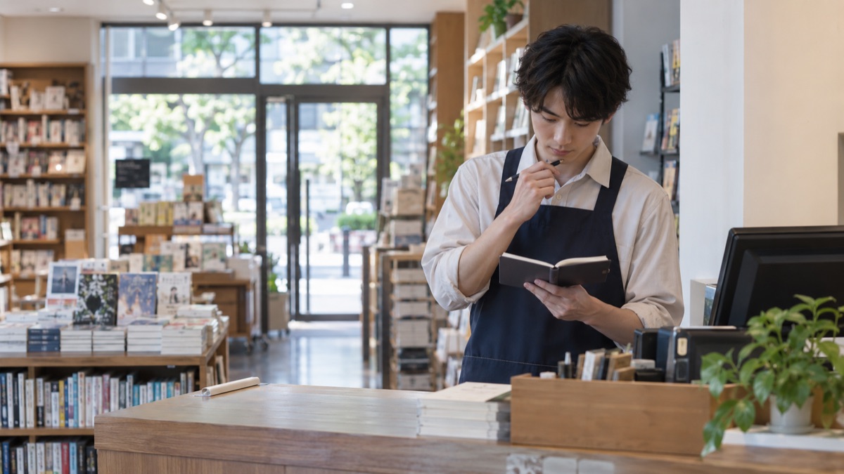 開店前の書店のカウンターでメモ帳を見返して準備する男性店員