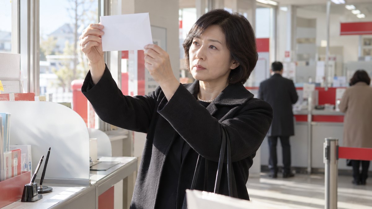 昼、郵便局の記帳台の前で、喪服の上にコートを着た50代の女性が、白い一重の封筒を光にかざして、中身が透けないかを確かめている。封筒や掲示の文字は読めない