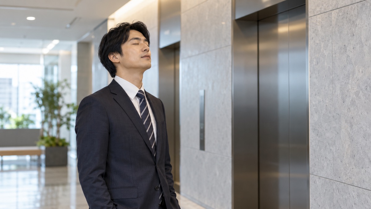 会議の前のエレベーターホールで、スーツの男性が目を閉じて息を長く吐いている