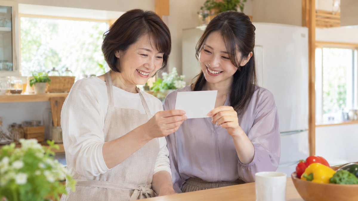 昼、実家の明るい台所のカウンターの前で、50代の母親と30代の娘が並んで立ち、白い無地のカードを手に取って見せ合いながら笑っている。カードに文字は無い
