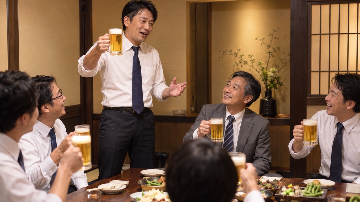 居酒屋の個室で、課長がジョッキを掲げて送別会の乾杯の音頭を取り、隣で主役がほほえんでいる