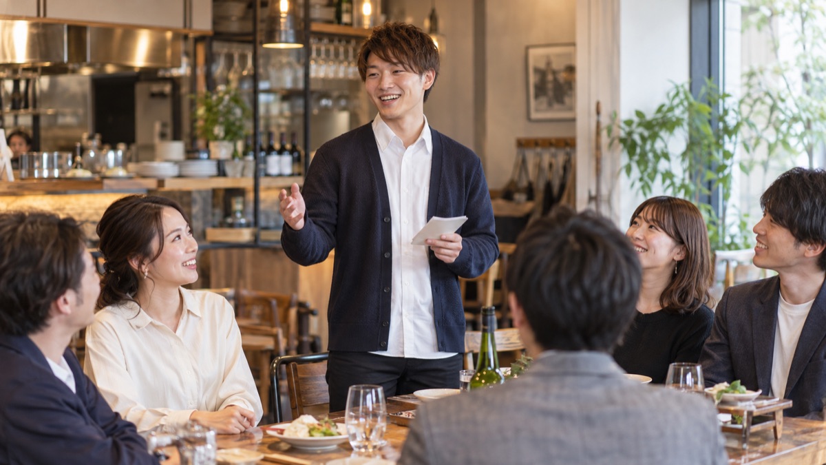 ビストロの店内中央で、若手の幹事が手元のメモを持って開会の挨拶をしている