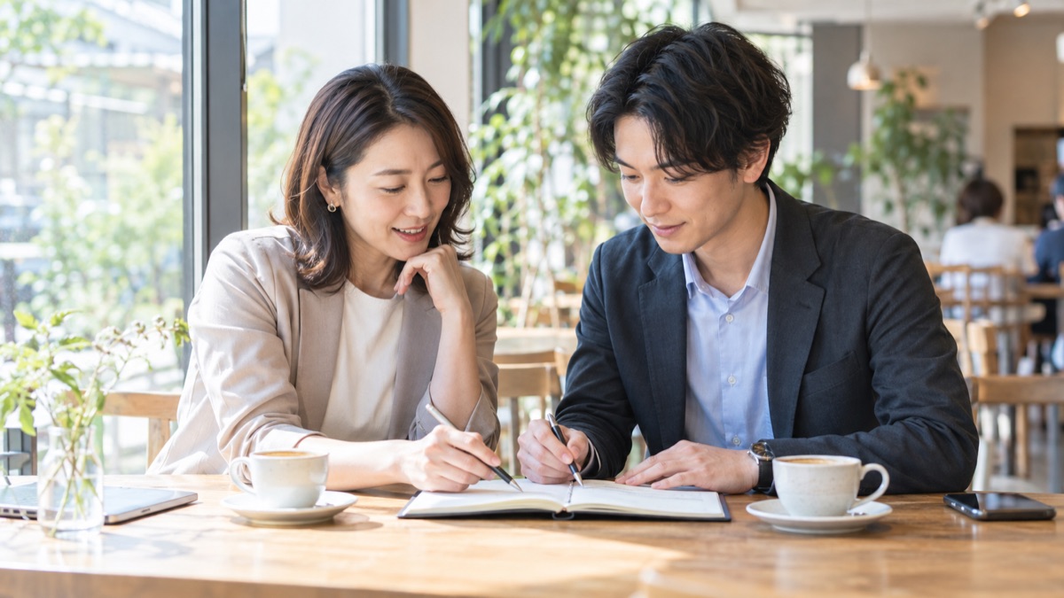 昼のカフェの窓際で、同僚2人が送別会で話す言葉をノートで3行に削っている