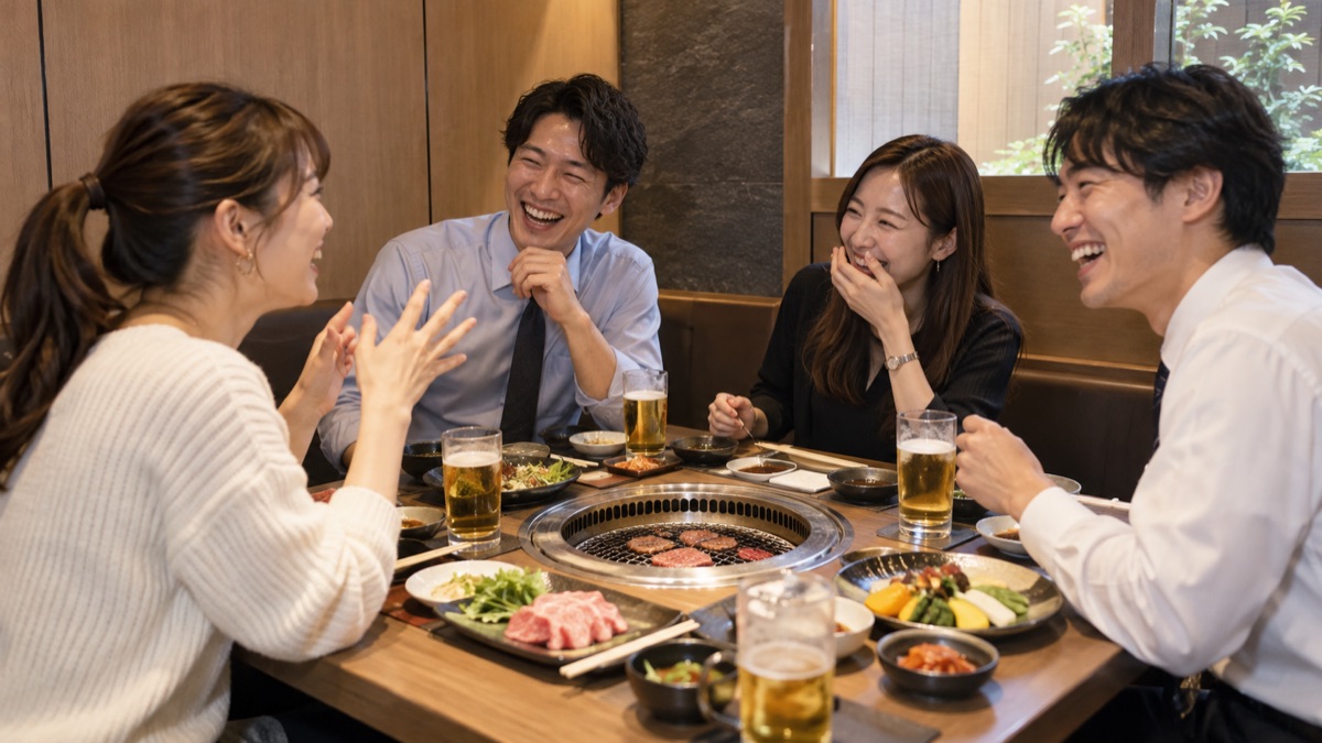 焼肉店の個室で、主役の女性が座ったまま少しおどけた一言を言い、同僚たちが笑っている