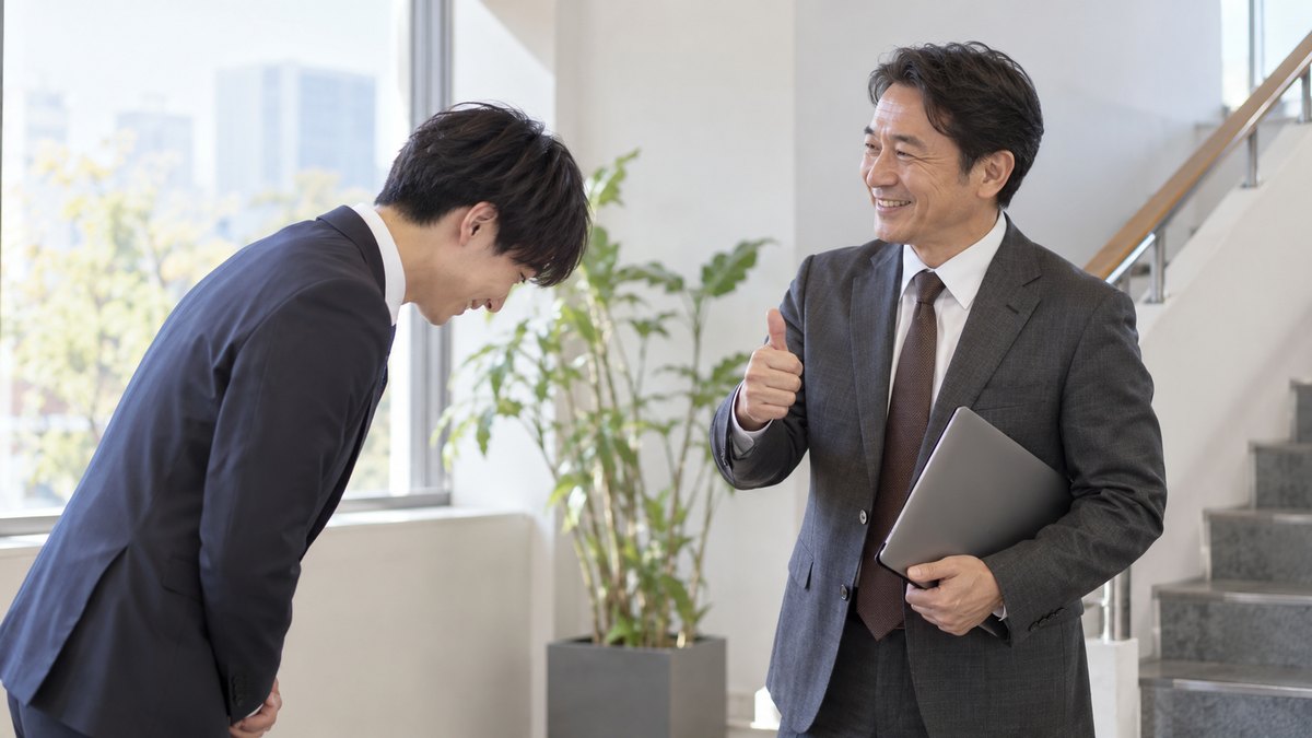 社内の階段の踊り場で、20代の男性幹事が40代の先輩社員に頭を下げてお願いし、先輩が笑顔で親指を立てて引き受けている