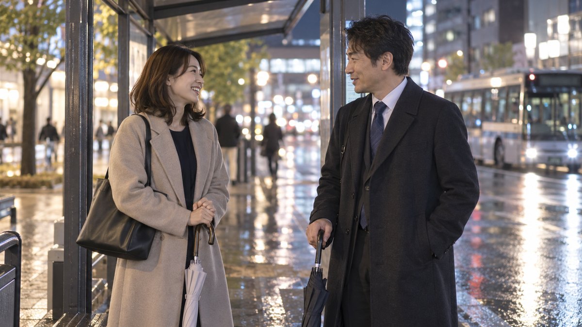 雨上がりの夜、駅前のバス停の屋根の下で、送別会帰りのコートを着た30代の女性と40代の男性が、閉じた傘を手に穏やかに話している