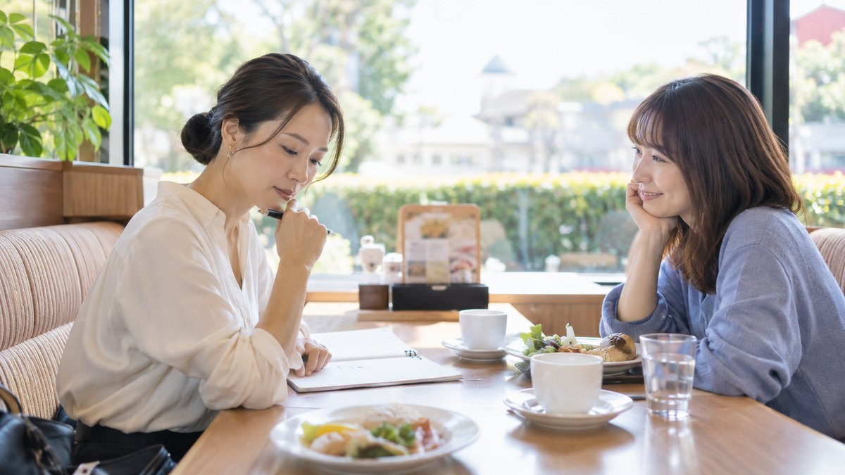 昼、ファミリーレストランの窓際の席で、30代の女性2人が向かい合って座り、片方がノートを開いて考え込み、もう片方がほおづえをついて笑顔で話を聞いている。メニューやノートの文字は読めない