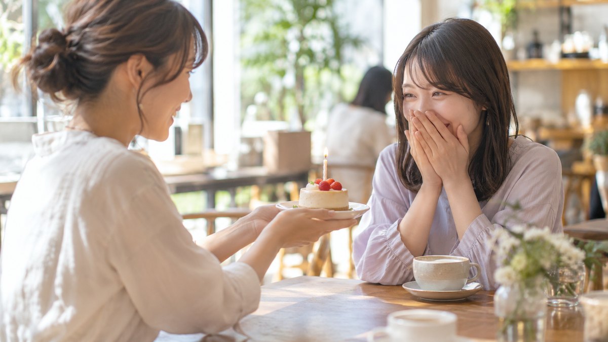 昼、明るいカフェのテーブルで、20代後半の女性が向かいの友人の前に、ろうそくを1本立てた小さなケーキをそっと差し出し、友人が両手で口元をおさえて喜んでいる