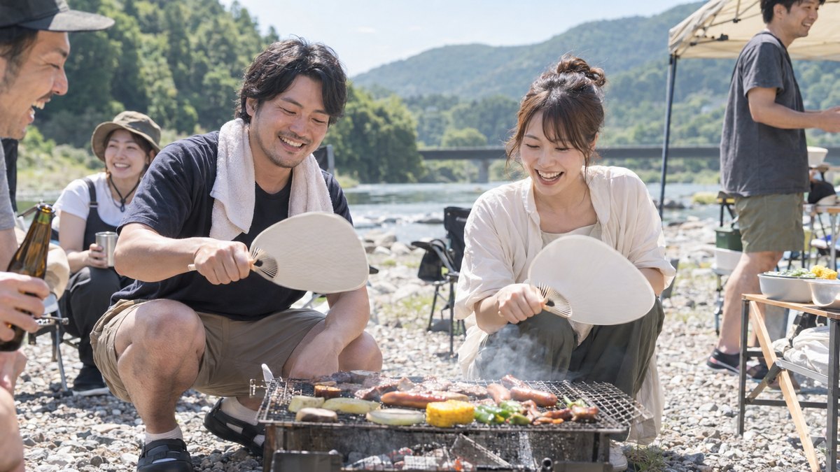 昼、河原のバーベキュー場で、30代の男性がしゃがんでうちわで炭をあおぎ、隣の30代の女性が笑いながら一緒にうちわを動かしている。まわりに友人たちが少し写る