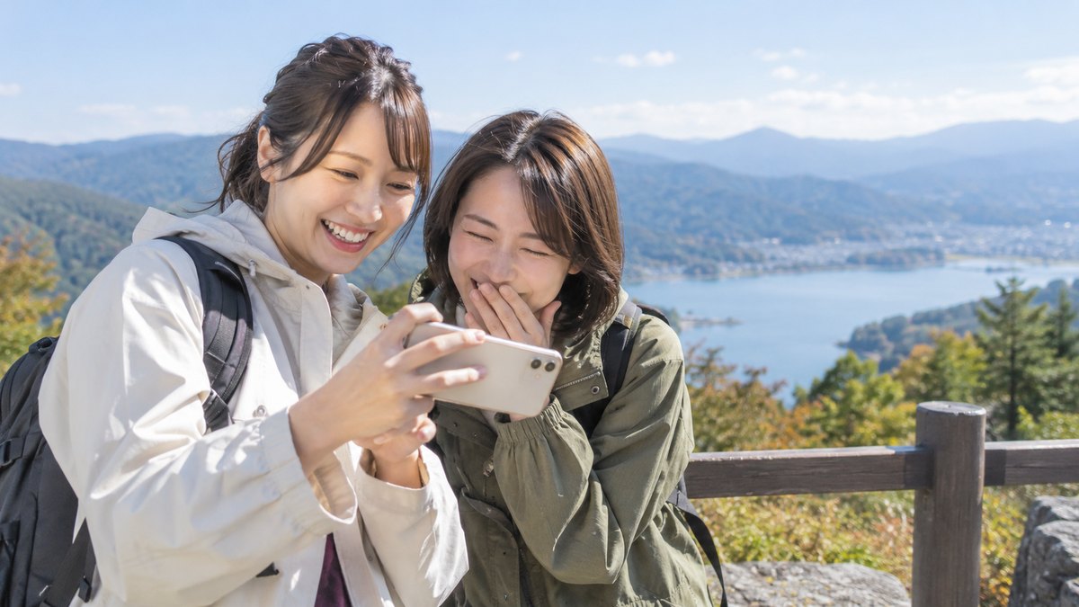 昼、旅行先の山の展望台で、20代後半の女性2人のうち一人がスマートフォンで写真を撮り、撮られた方が目をつぶってしまった画面を二人でのぞき込んで笑っている。画面は見えない
