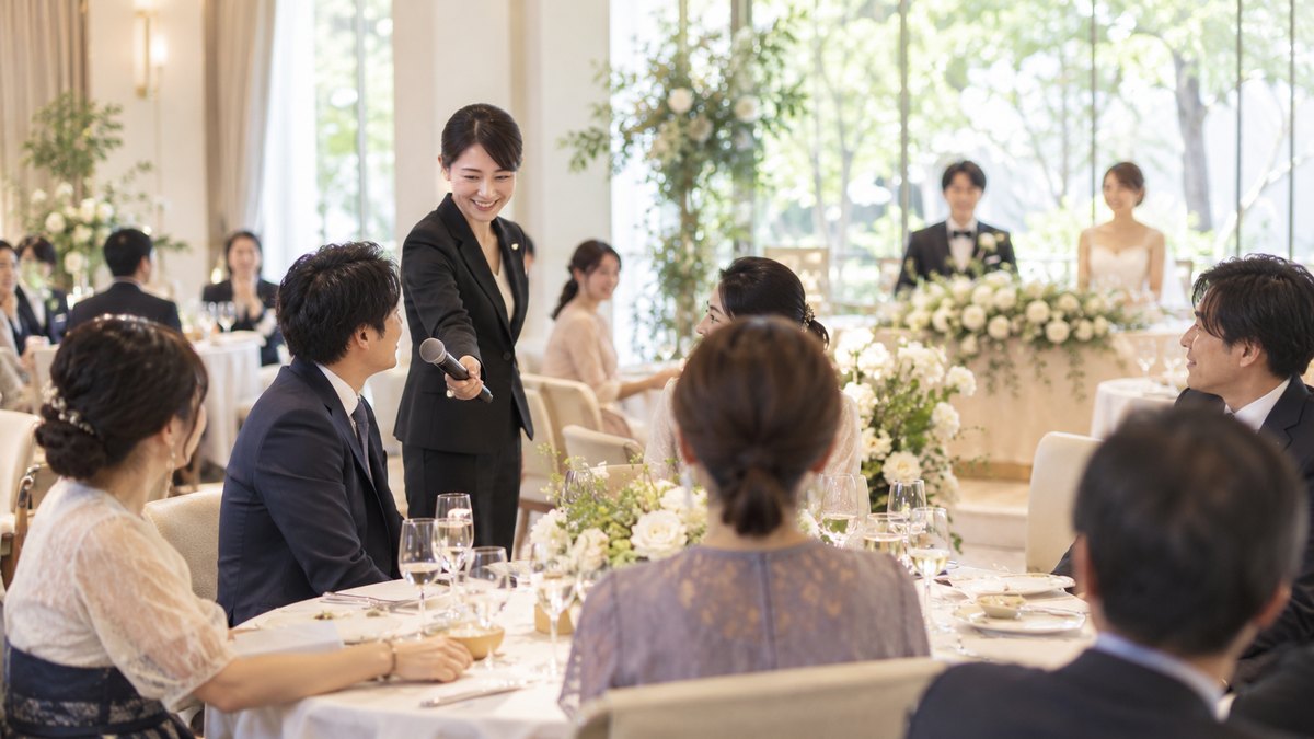 昼、明るいレストランウエディングの会場で、黒いスーツの女性司会者がワイヤレスマイクを持ってゲストの円卓を回り、座ったままの30代の男性ゲストにマイクを向けている。顔は小さく写る