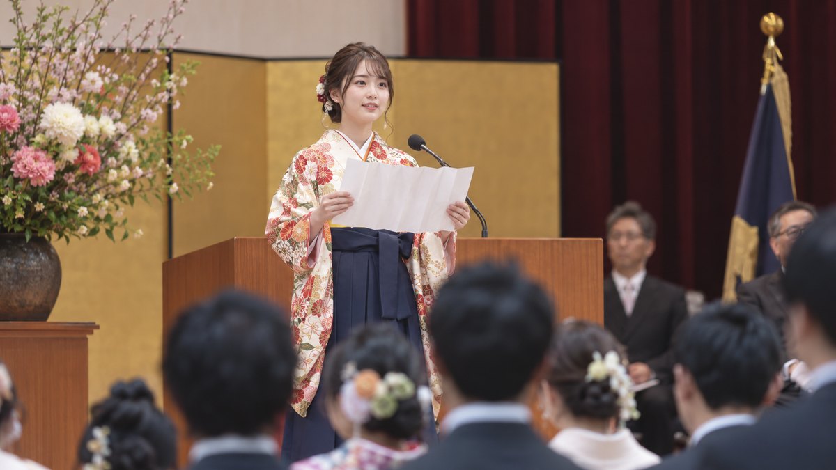 大学の卒業式の講堂で、袴姿の女性が演台の前に立ち、蛇腹折りの答辞の紙を広げて前を向いて読んでいる
