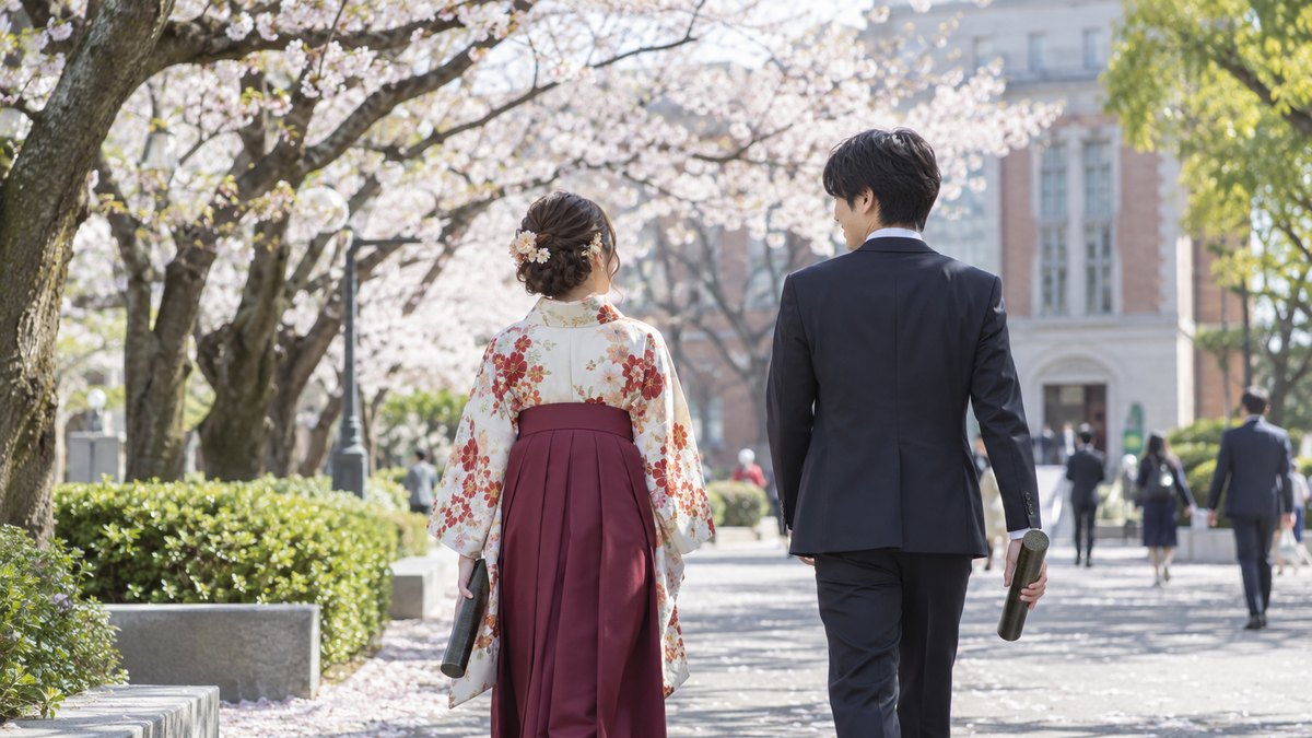 卒業式を終えた春の大学キャンパスの並木道を、袴姿の女性とスーツ姿の男性が、卒業証書の筒を手に並んで歩いていく後ろ姿