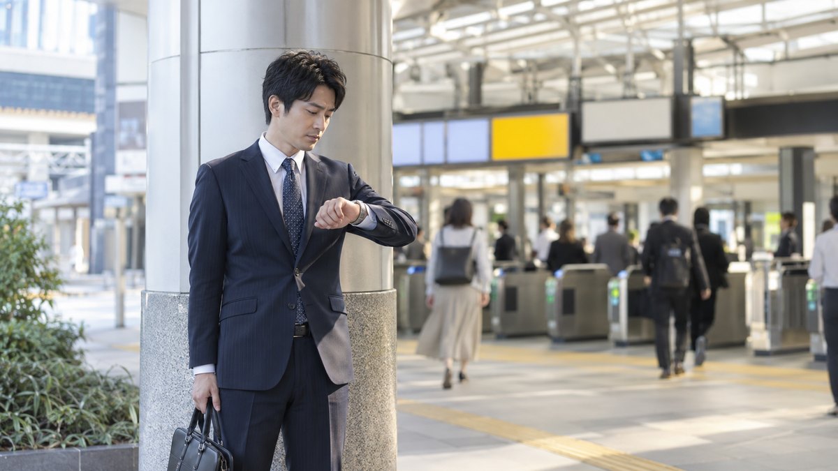 平日の朝、駅の改札の外の柱のそばで、スーツ姿の30代の男性が待ち合わせの時間よりずっと早く着き、腕時計を確かめながら一人で立っている。案内表示の文字は読めない