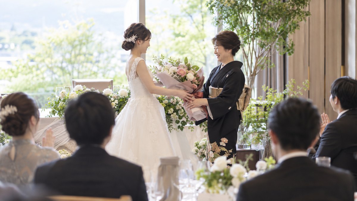 昼、披露宴会場の前方で、白いドレスの新婦が留袖の60代の女性に大きな花束を手渡し、女性が両手で受け取ってほほえんでいる。顔は小さく写る