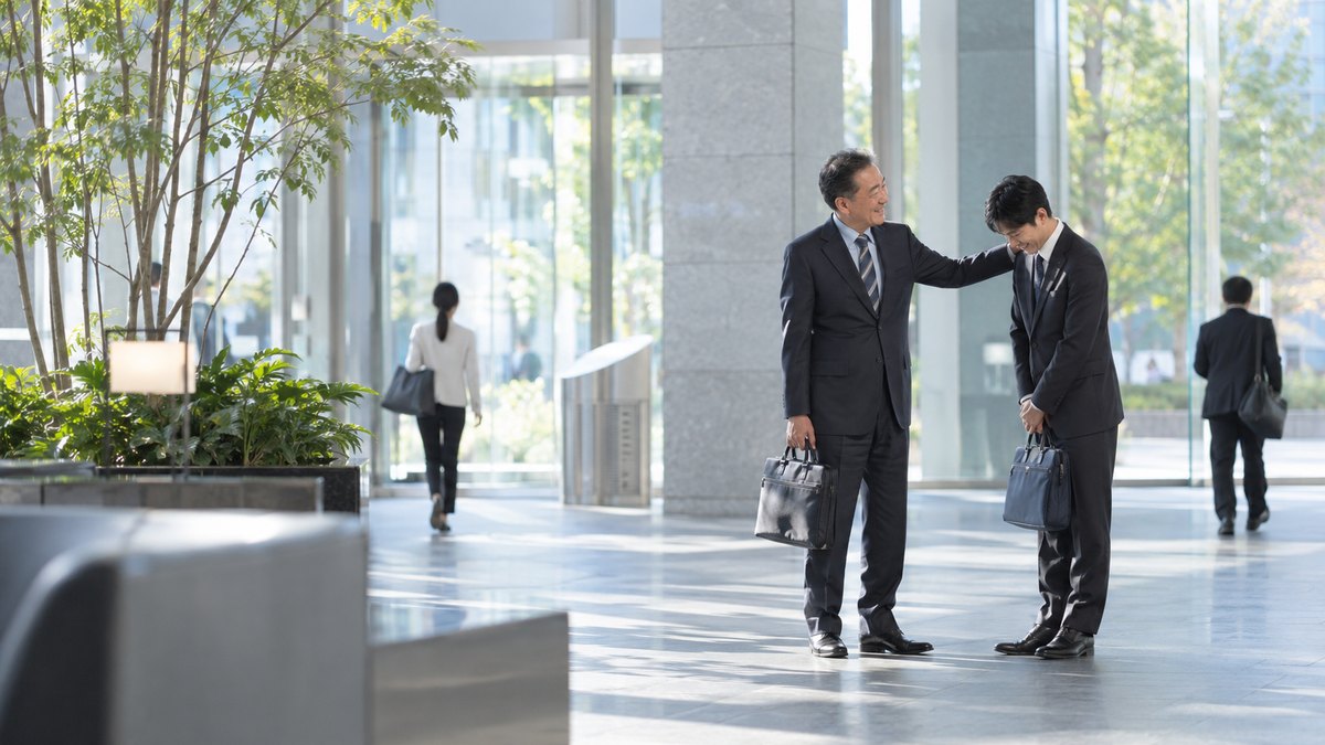 昼、オフィスビルの1階の明るいロビーで、スーツ姿の50代の男性上司が、30代の男性部下の肩に軽く手を置いて笑顔で声をかけ、部下が照れたように頭を下げている。顔は小さく写る。看板の文字は読めない