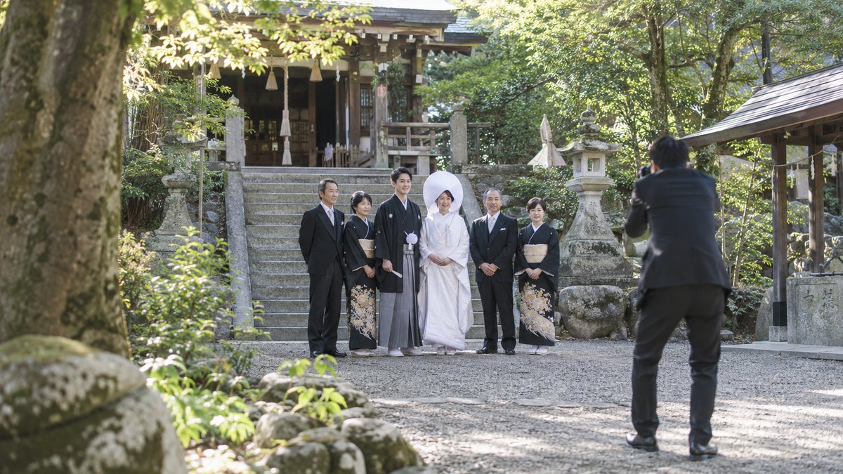 昼、小さな神社の境内の石段の前で、白無垢の新婦と紋付袴の新郎を真ん中に、礼服の家族6人ほどが並び、カメラマンに写真を撮ってもらっている。顔は小さく写る。文字は写らない