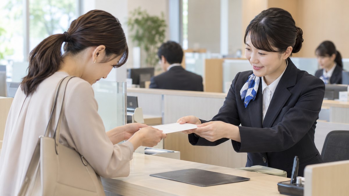 昼、銀行の窓口で、30代の女性が窓口の女性行員から、口を閉じた白い無地の封筒を両手で受け取り、軽く頭を下げている。封筒の中身やお札は見えない。窓口の掲示の文字は読めない