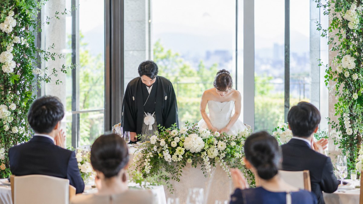 昼、明るい披露宴会場の高砂の席で、白いドレスの新婦と紋付袴の新郎が、軽く頭を下げてから椅子に腰を下ろそうとしている。奥の窓から光が差す。顔は小さく写る