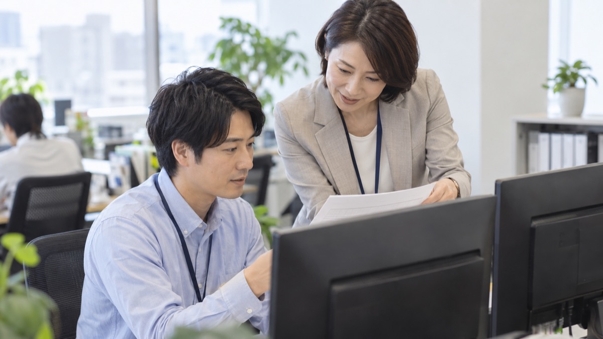 デスクで女性上司が部下の横にかがみ、紙のチェック表を一緒に確認する