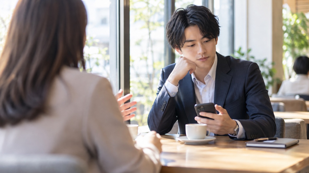 カフェで同僚の話を聞きながら、手元のスマートフォンに目をやる男性