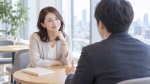 職場の打ち合わせテーブルで、向かいの同僚の話をうなずきながら聞く女性会社員