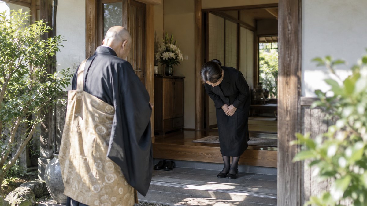 朝、古い一軒家の玄関で、喪服の50代の女性が、到着した袈裟姿の70代の僧侶を迎え、上がりかまちの手前で深く頭を下げている。顔は小さく写る。文字は無い