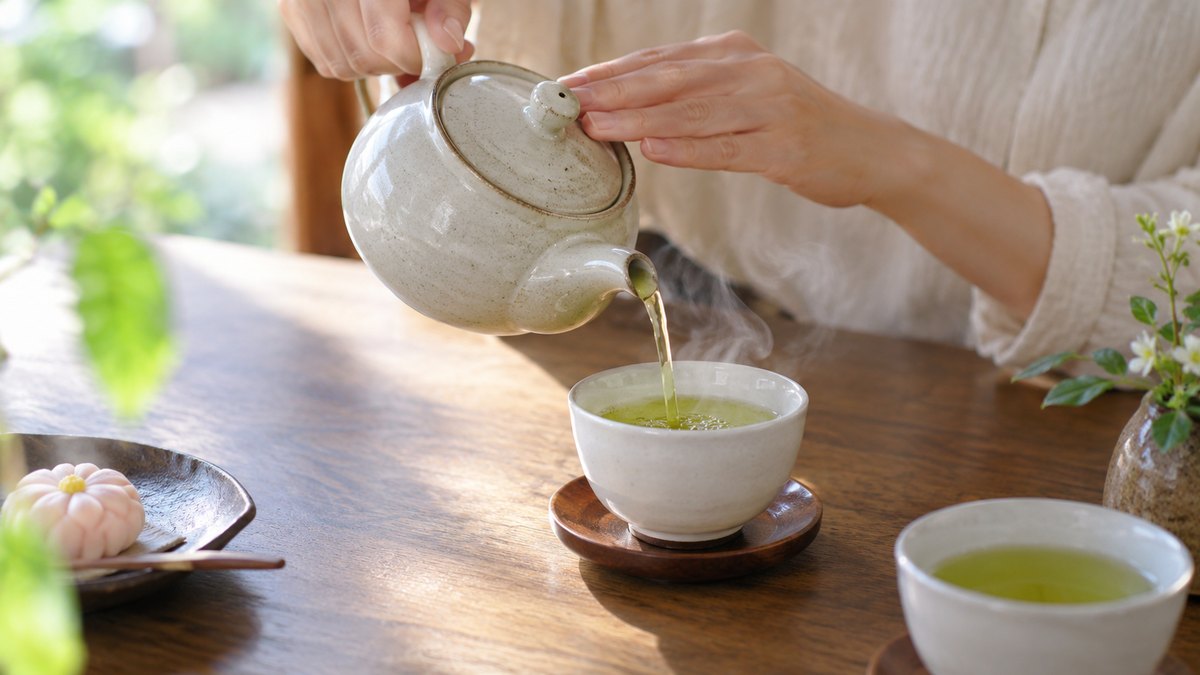 昼、木のテーブルの上で、急須から白い湯のみに緑茶を静かに注いでいる手元のアップ。湯気が立っている。顔は写らない。文字は無い