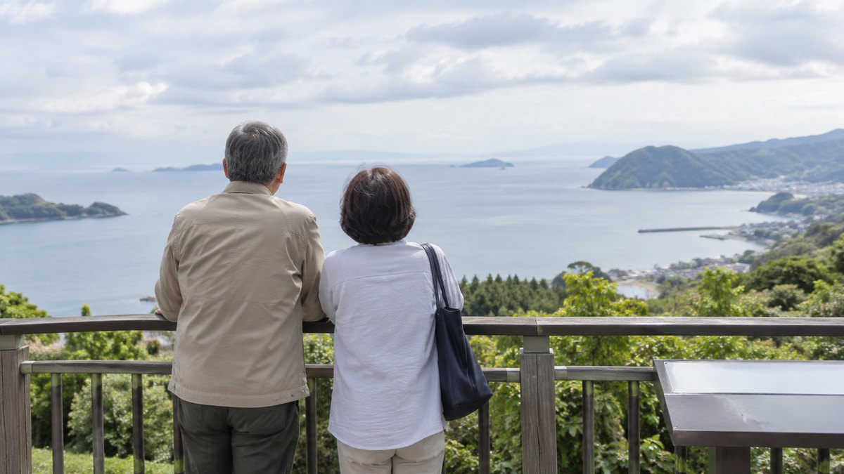 曇りの昼、海の見える高台の展望台で、普段着の60代の夫婦が手すりのそばに並んで立ち、遠くの海を静かに眺めている後ろ姿。看板の文字は無い