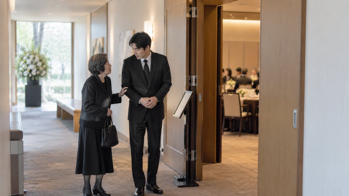 昼、斎場の会食室の入口の廊下で、喪服の70代の女性が30代の男性の腕に軽く手を添えて小声で何かを頼み、男性が少し驚いた顔でうなずいている。顔は小さく写る。案内板の文字は読めない