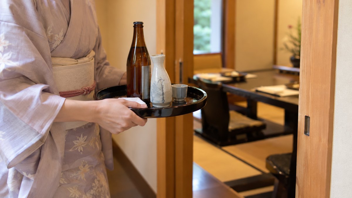 昼、和食店の個室の入口で、着物の仲居が盆にのせた瓶の烏龍茶と日本酒の徳利を運んでいる手元。顔は写らない。ラベルや品書きの文字は読めない