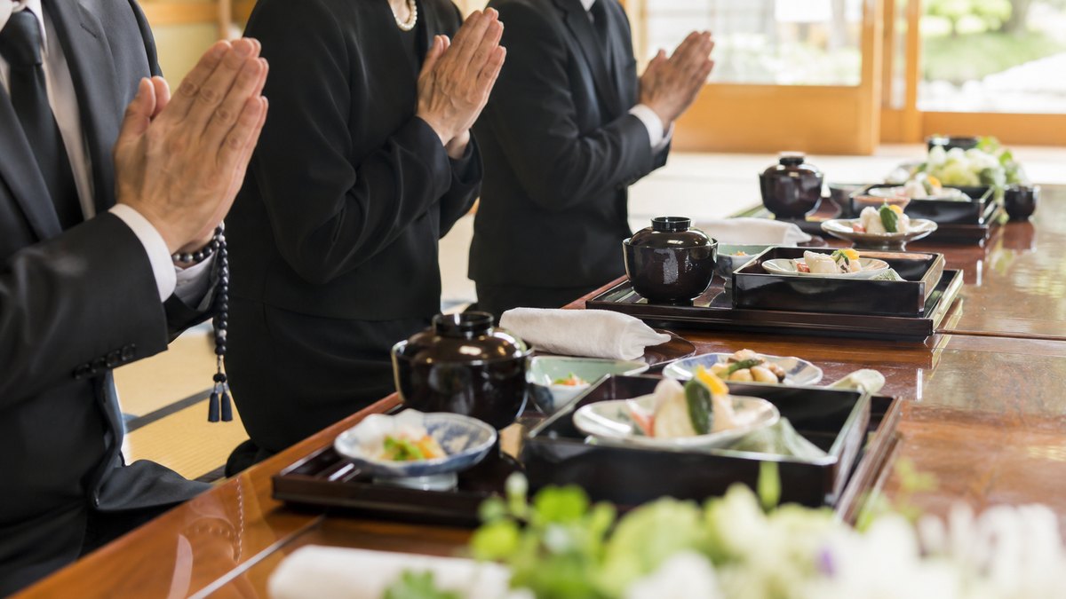 昼、寺の客殿の座卓で、喪服の親族数人が料理を前に静かに手を合わせている手元。顔・僧侶・祭壇は写らない。文字は無い
