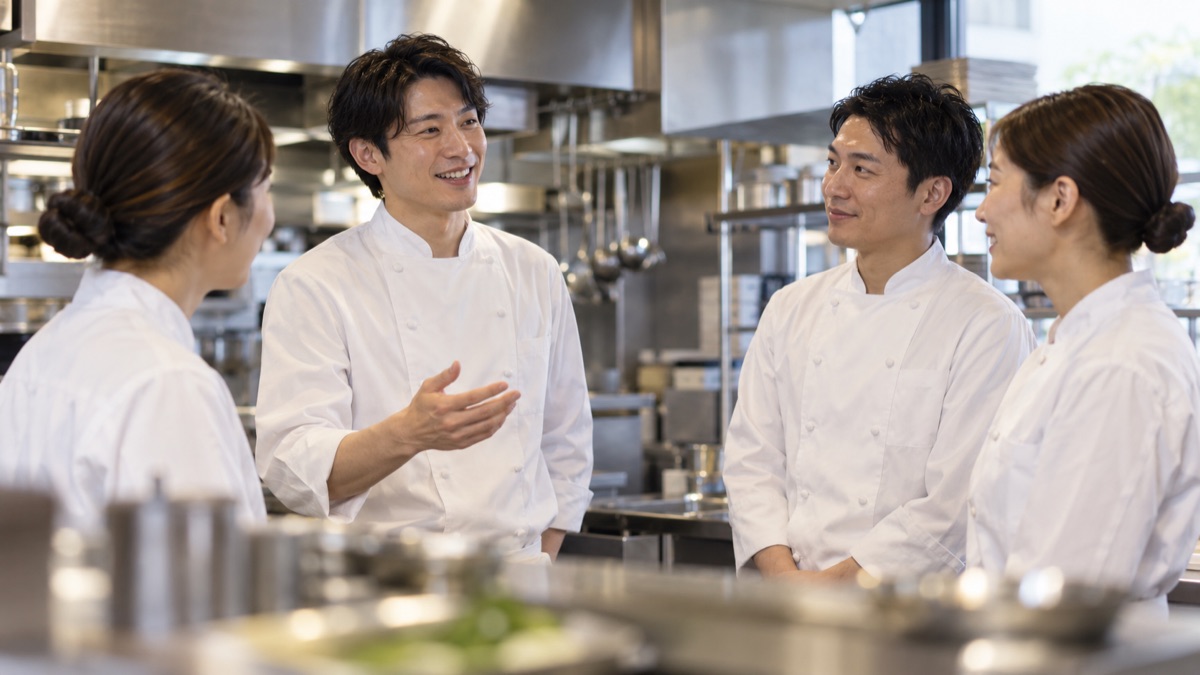開店前の厨房で料理人たちが短く声をかけ合う朝の場面