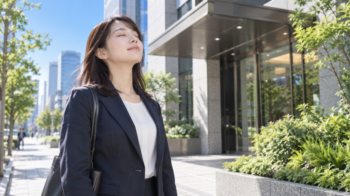 会社のエントランス前で目を閉じて深呼吸する新入社員の女性
