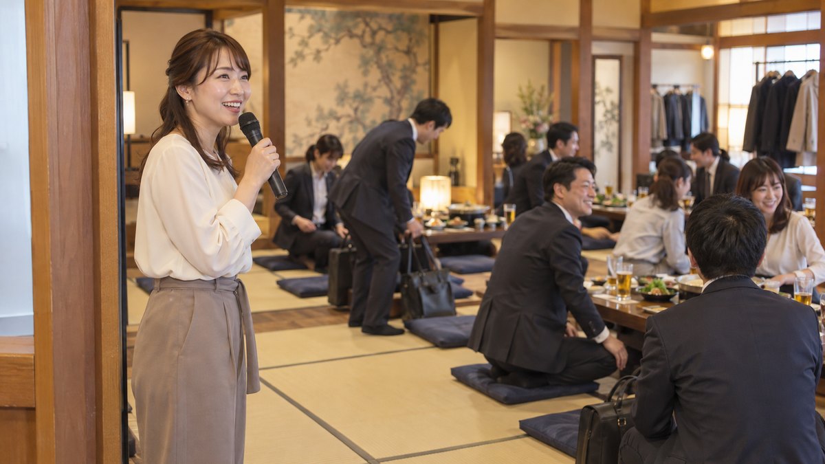 居酒屋の広い座敷の入口近くで、30代の幹事の女性がマイクを手に座敷を見渡し、到着した社員たちが座布団に座り始めている