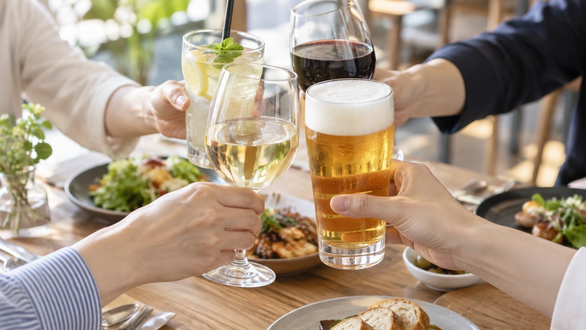 居酒屋ではない明るいカフェレストランの木のテーブルの上で、何人もの手がそれぞれのグラスを一斉に持ち上げている手元のアップ。顔は写らない