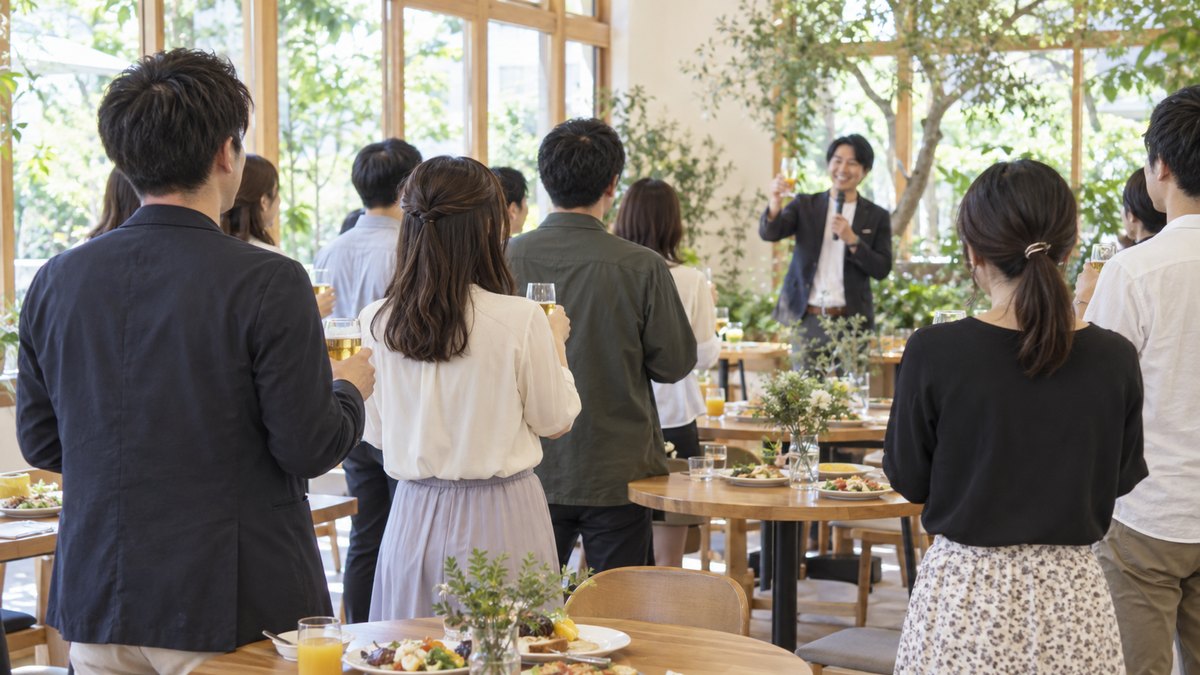 昼の明るい郊外のガーデンレストランの室内で、緑の多い窓辺に木の丸テーブルが並び、カジュアルな服装の社員たちが立ったまま前方の司会者を見て、テーブルのグラスに手を伸ばし始めている後ろ姿。金屏風も高層ビルの窓も無い