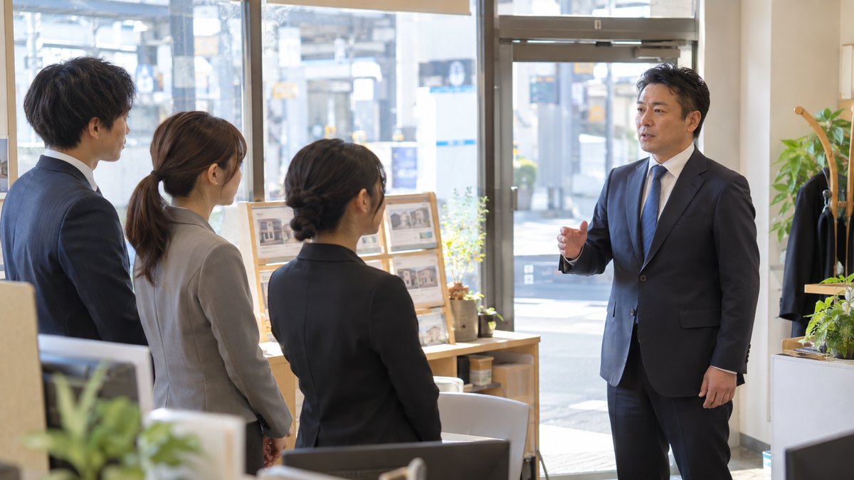 1月の朝、駅前の小さな不動産会社の店内で、スーツ姿の40代の男性店長が開店前に立ったまま、3人のスタッフに向かって短く話している。張り紙や画面の文字は見えない