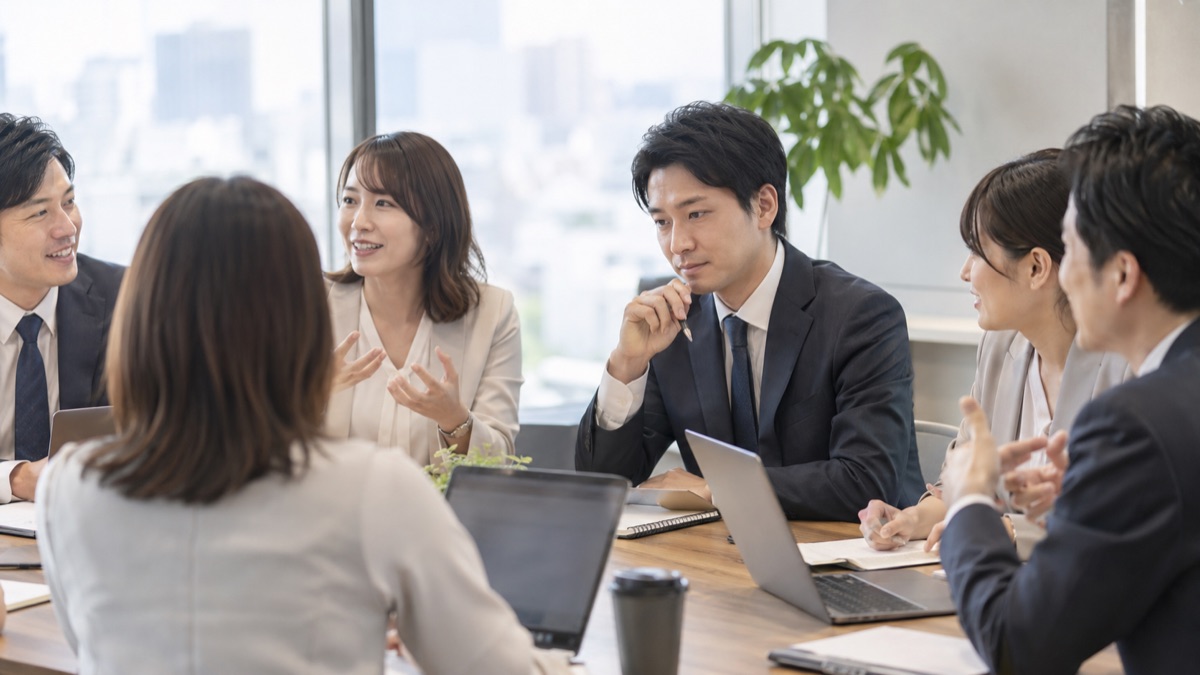 会議テーブルで同僚が話す中、ペンを握ったまま言い出せずにいる男性社員