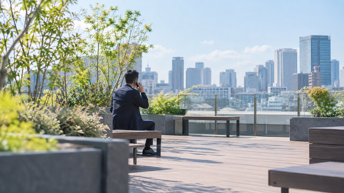 昼、オフィスビルの屋上の休憩スペースのベンチで、40代の男性会社員がスマホを耳に当て、穏やかな表情で電話をしている遠景。顔ははっきり写らない。文字は無い