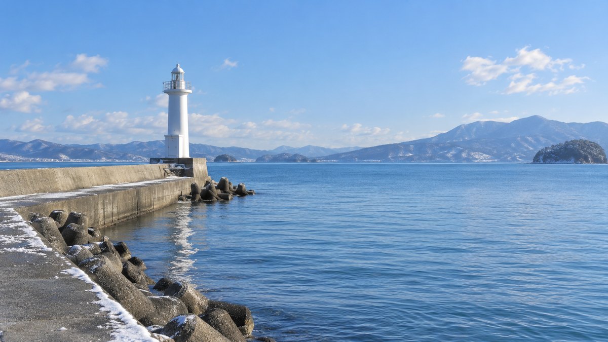 冬の晴れた日の、港の防波堤の先にある白い灯台と穏やかな海の遠景。人物はいない