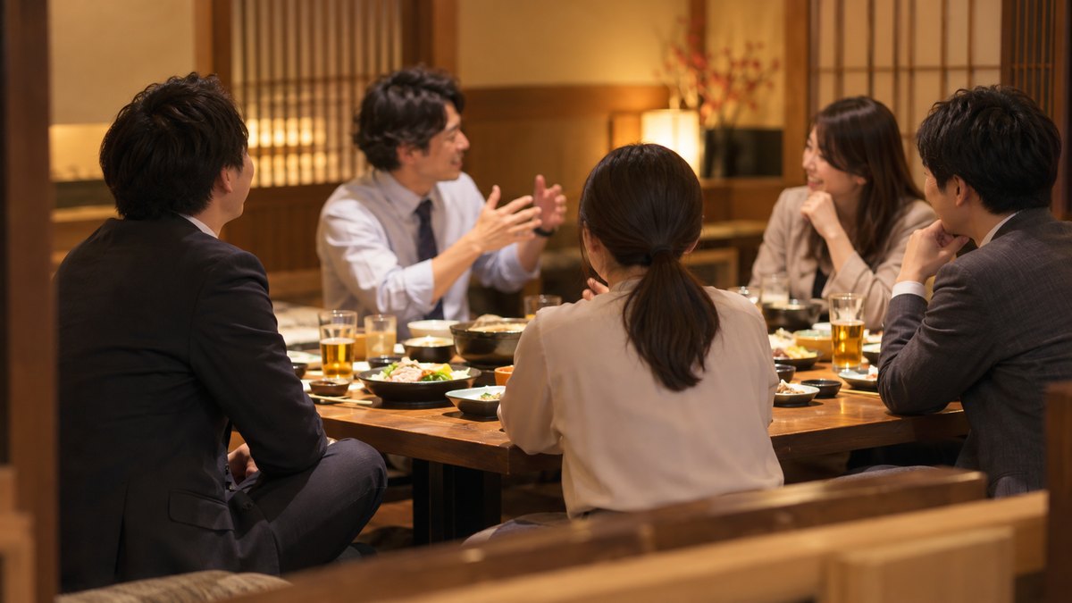夜、居酒屋の座敷の長いテーブルで、会社の同僚5人が食事をしている中、端の席の30代の男性会社員が中心で話す同僚のほうを向いて笑顔でうなずいている遠景。顔ははっきり写らない。メニューや店の文字は見えない