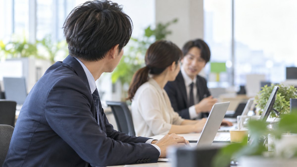 午前、オフィスの島型の席で、30代の男性会社員が自分のパソコンに向かったまま、隣の席の同僚たちが話しているほうを少しだけ見ている横からの近景。パソコンの画面は見えない角度。顔ははっきり写らない。文字は無い