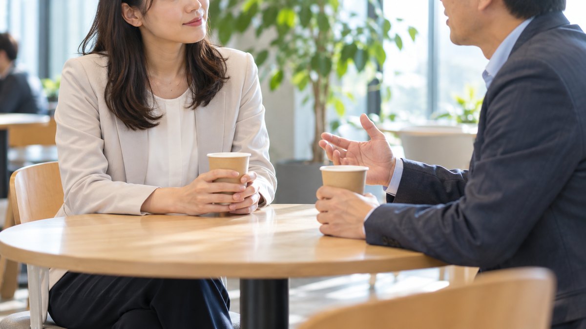 昼、社内の休憩スペースの丸テーブルで、30代の女性会社員と40代の男性の同僚が紙コップを手に向かい合い、女性がうなずきながら相手の話を聞いている近景。顔ははっきり写らない。文字は無い