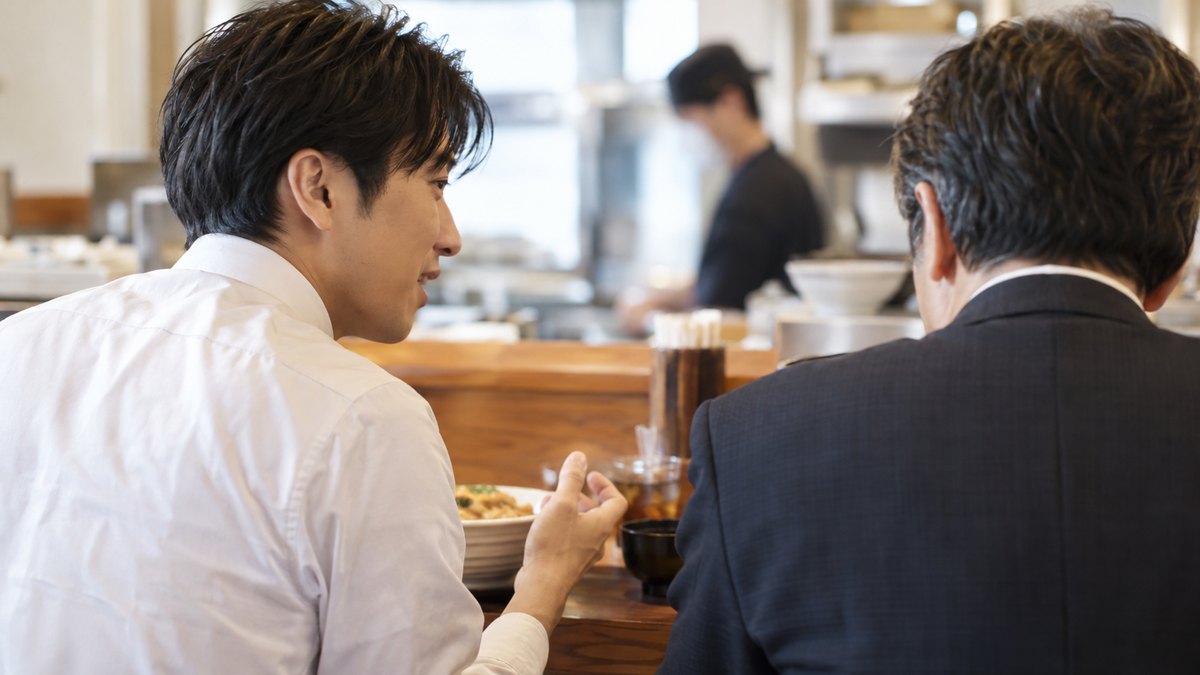 昼、会社の近くの定食屋のカウンター席で、30代の男性会社員が隣に座る同じ職場の40代の男性に、顔を向けて話しかけようとしている肩から上の近景。顔ははっきり写らない。メニューや店の文字は見えない