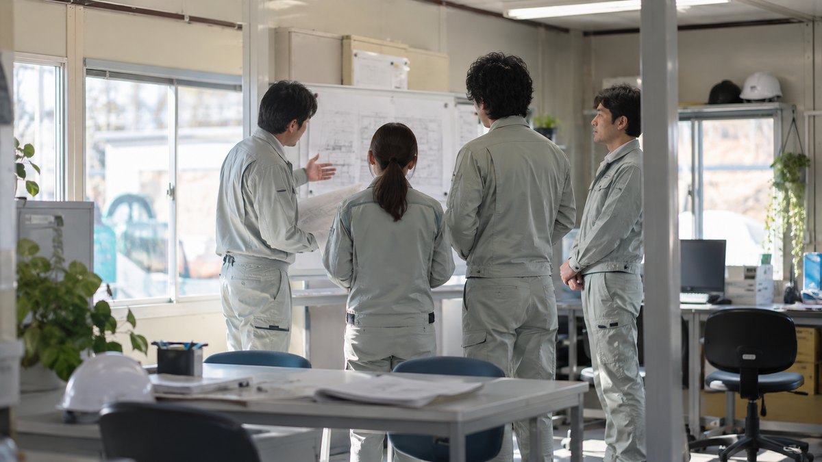朝、建設会社の現場事務所で、作業着の4人が立って短い打ち合わせをしていて、端の30代の男性が話す人のほうを見てうなずいている遠景。顔ははっきり写らない。図面や掲示の文字は読めない