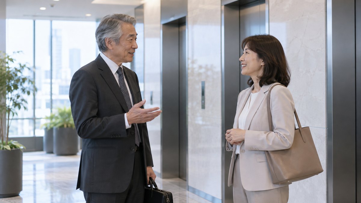オフィスビルのエレベーターホールで、60代の男性が他部署の50代の女性と向き合い、短く立ち話をしている