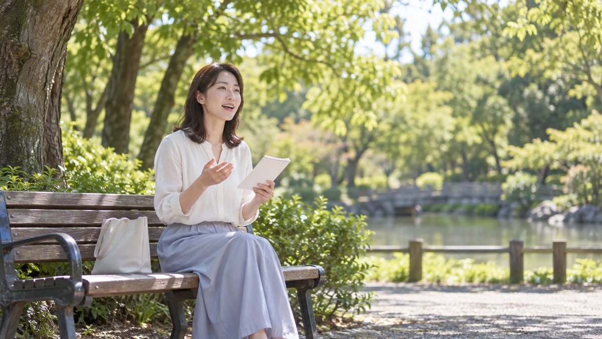 週末の午後、公園の木かげのベンチで、30代の女性が小さなメモを片手に持ち、前を向いて声に出して話す練習をしている遠景。メモの文字は見えない