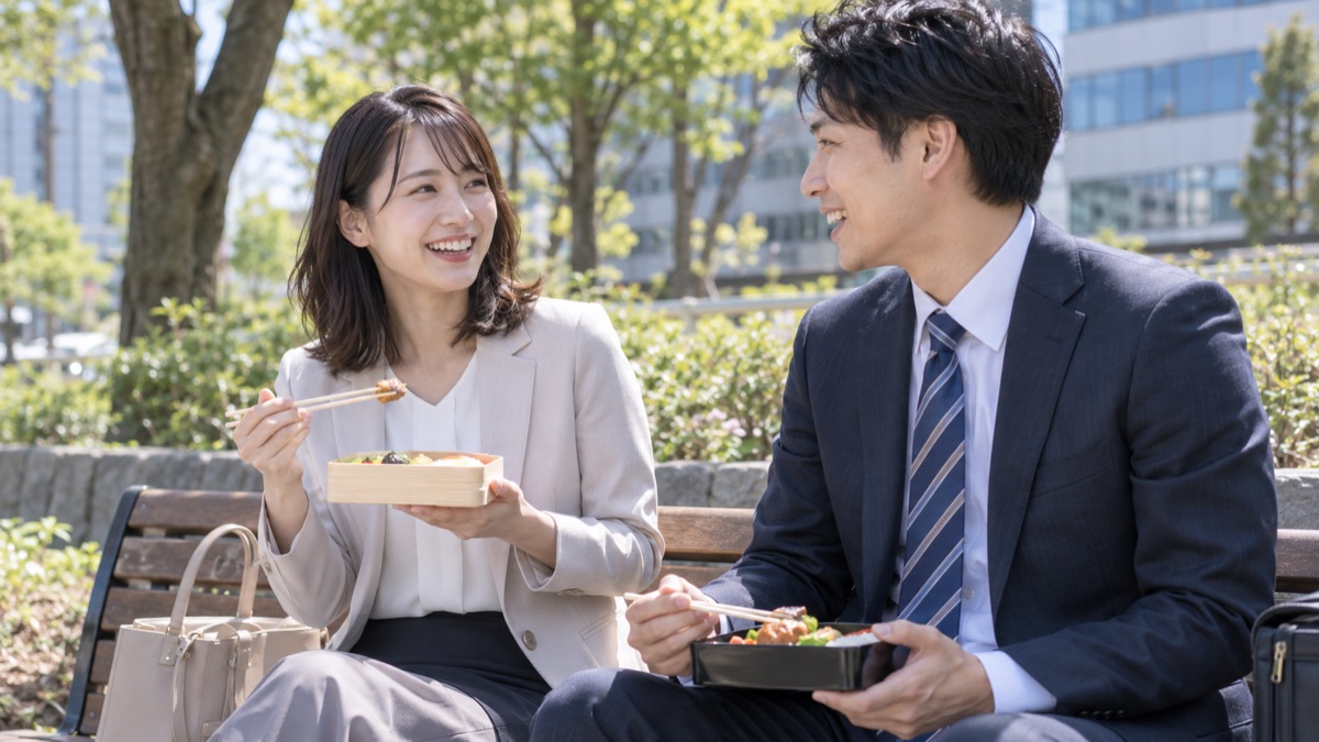 昼休みの公園のベンチで、同僚と弁当を食べながら楽しそうに話す女性会社員